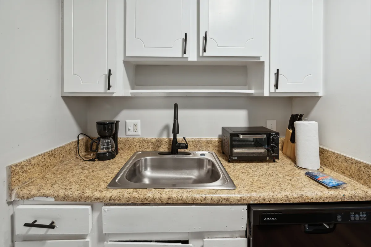 A cozy kitchen scene with a sink, coffee maker, toaster oven, and paper towels on the counter. The warm, inviting setup hints at morning routines, casual breakfasts, or preparing snacks.