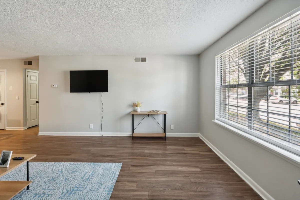 This image displays a welcoming living area featuring a TV mounted to the wall, a small table adorned with a floral arrangement, and a large window with blinds that lets in natural light. The room has a comfortable and open feel, suggesting a relaxing environment ideal for either unwinding or socializing.