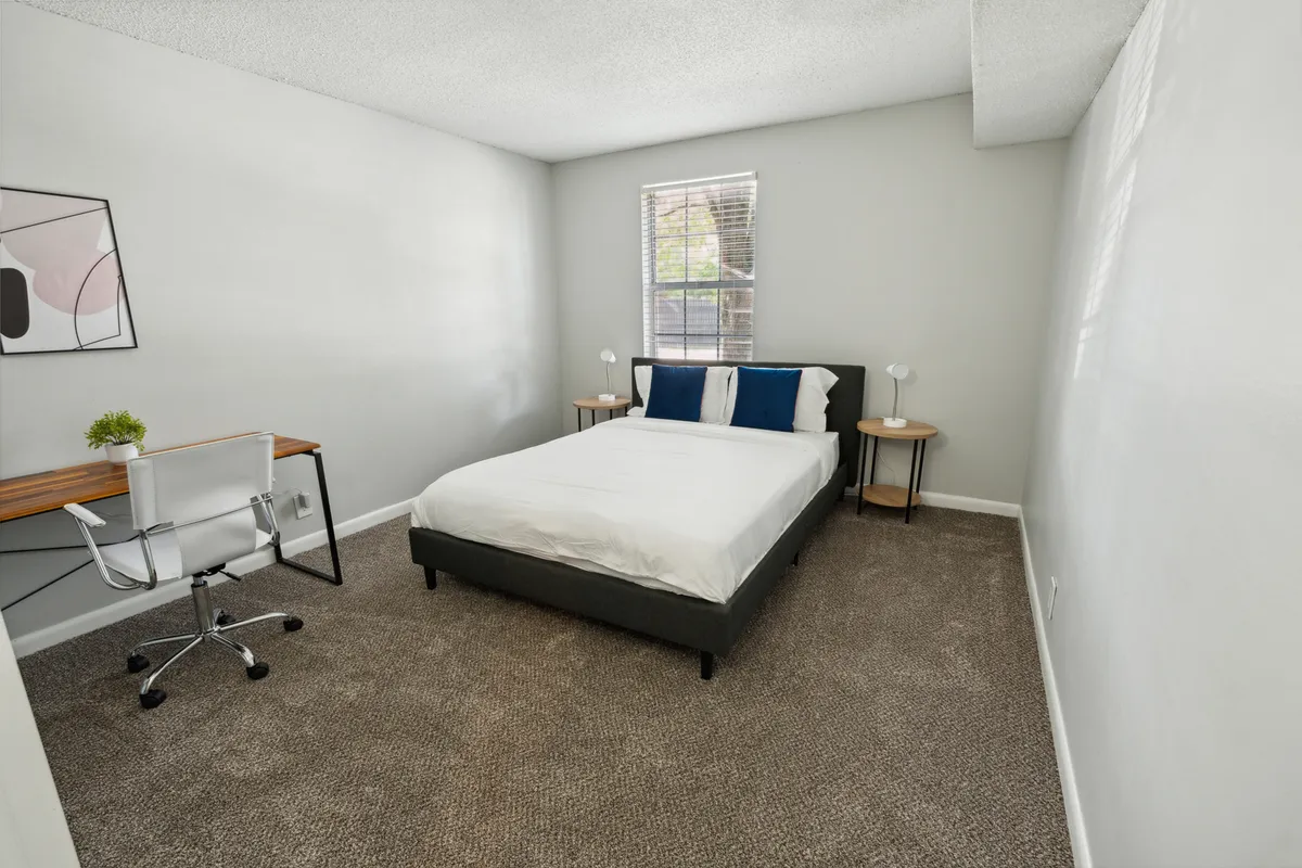 A tranquil bedroom featuring a plush bed dressed in white bedding, accented by a desk and chair near a window letting in abundant natural light. Abstract wall art adds a touch of sophistication, while the overall atmosphere is calm and restful—ideal for unwinding or catching up on work.
