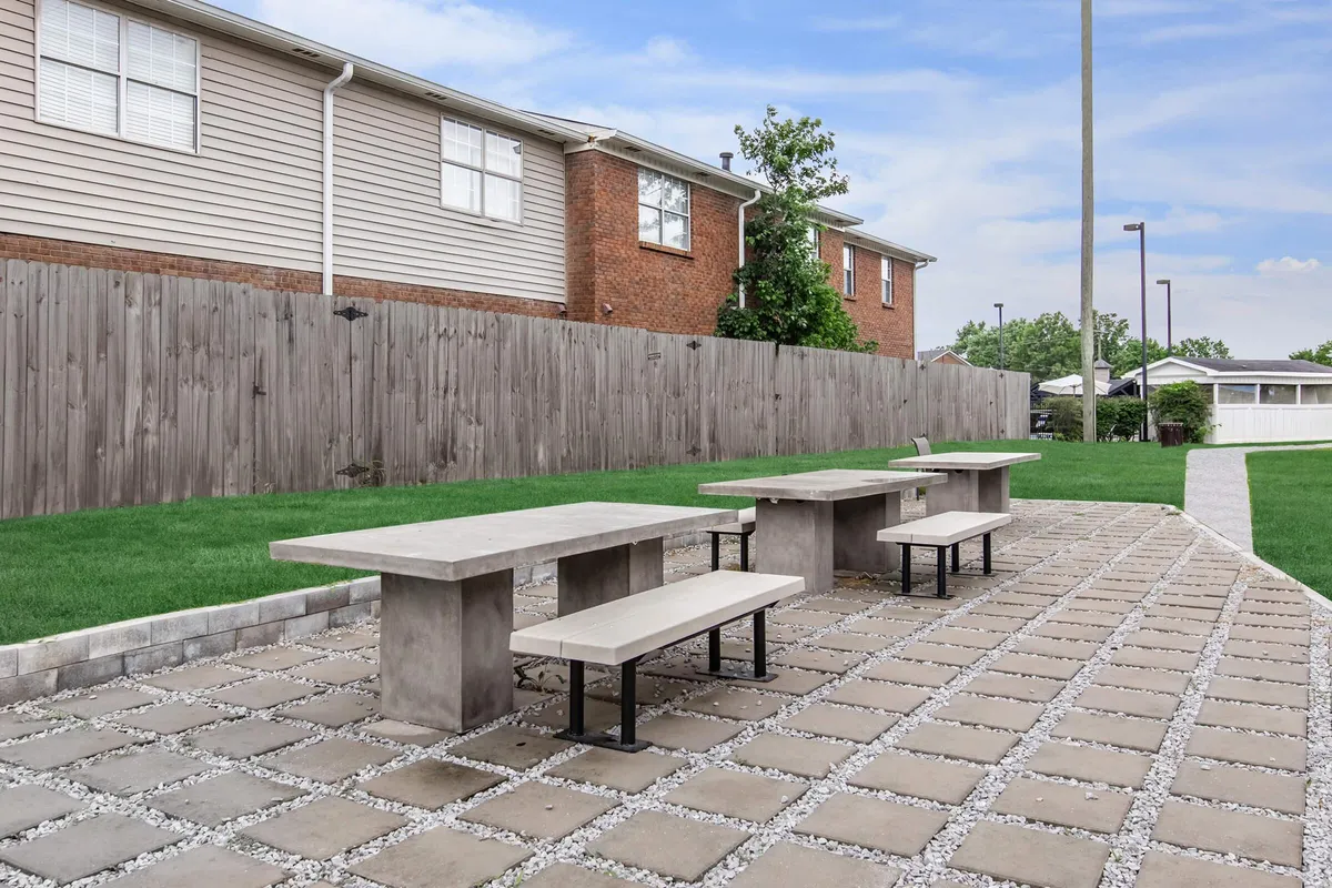 An inviting communal outdoor area featuring concrete tables, benches, landscaped grass, and a wooden fence—ideal for social gatherings, outdoor dining, or casual relaxation.