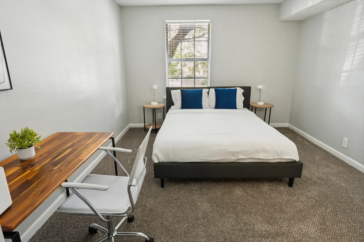 The image showcases a serene and inviting bedroom space with a neatly made bed adorned with blue and white accent pillows, a wooden desk with a white chair set up as a potential workspace, and warm table lamps adding a cozy glow. Sunlight gently filters into the room, enhancing the tranquil atmosphere and highlighting the modern, stylish decor.