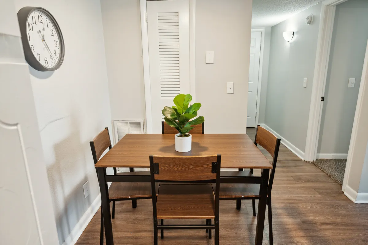 A bright and cozy dining area featuring a round table, comfortable chairs, a decorative plant, and a large wall clock. The arrangement is inviting, hinting at intimate meals, friendly gatherings, or quiet moments spent working at the table.