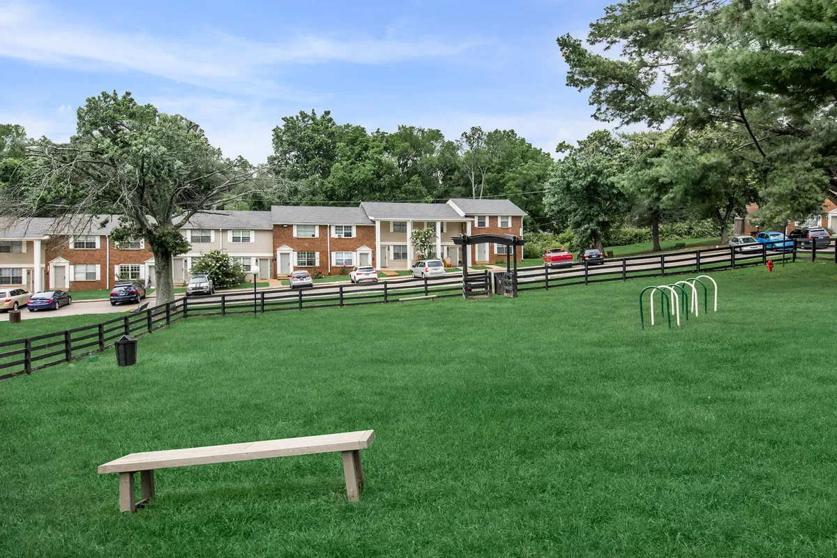 A wide green lawn dotted with playground equipment, benches, and a row of houses in the background. The mood is communal and inviting, with space for family gatherings, picnics, and outdoor relaxation.